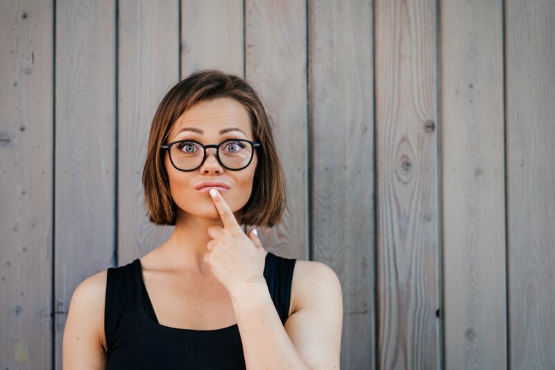 Pretty young woman in glasses dressed casual standing against wooden wall looking thoughtful, touching her lips with index finger, making decision. You have to choose.
